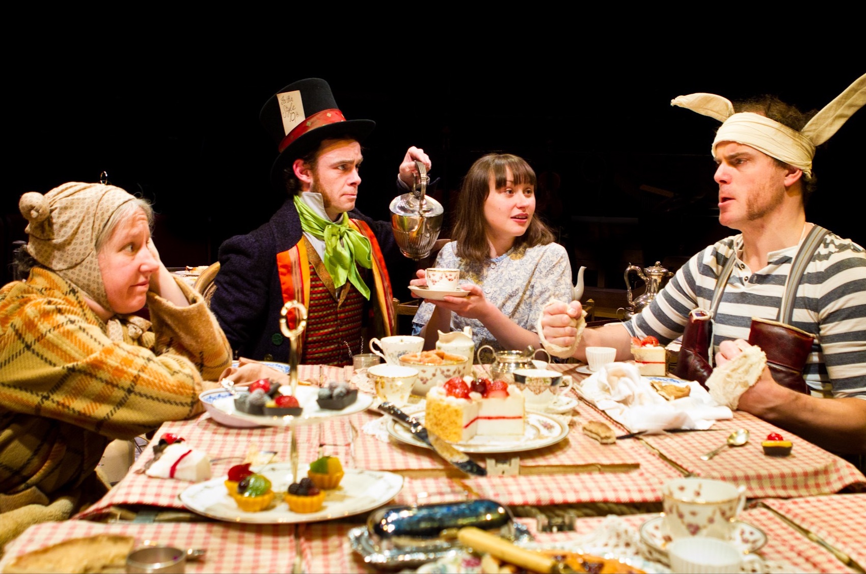 The Mad Hatter's tea party. We see four characters set around a table full of cups, saucers, tea and cakes. A dormouse in a dressing gown (played by a woman) looks fed up. The Mad hatter is pouring a cup of tea for Alice. All three are looking at the Mad March Hare (played by a man) who has big ears and fists ready for a punch up.