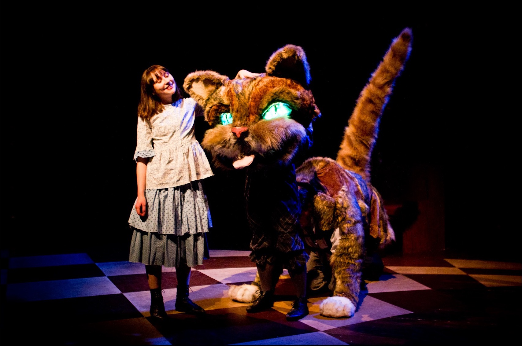 Alice stands on a chequerboard floor, next to a huge cat. She is smiling and stroking its head. The cat is a puppet operated by three people. It has bright green eyes and is sticking it's tongue out.