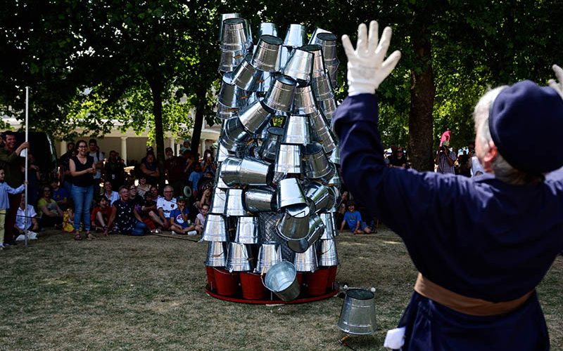 A sunny day in a park, with parched grass and a line of trees in the background. We see a large tower of buckets is beginning to fall. A person in a blue uniform is putting their hands in the air in alarm in the foreground. We see a large crowd in the background.