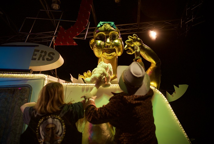 A large, golden baby head is sticking out the top of a caravan and there are two figures below it.