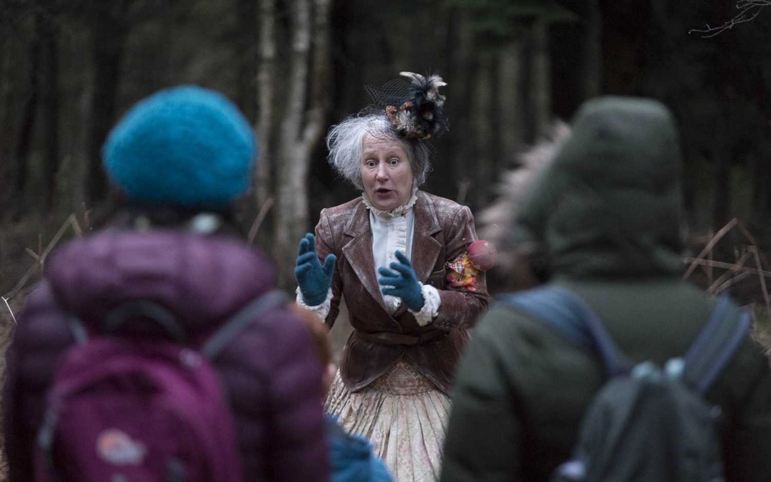 We see a woman with grey hair, wearing victorian clothes and a small top hat perched on one side of her head. She is appealing to us, the audience. Two members of the audience wearing winter clothes are in the foreground.