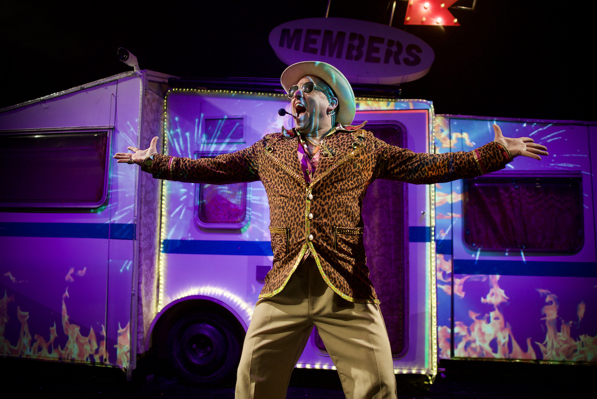 A man, wearing a leopard skin jacket and with outstretched arms. He looks excited and is shouting. Behind him is a caravan with bright lights and the image of flames projected onto it.