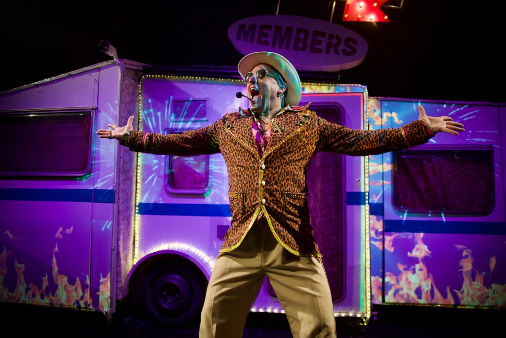 A man, wearing a leopard skin jacket and with outstretched arms. He looks excited and is shouting. Behind him is a caravan with bright lights and the image of flames projected onto it.